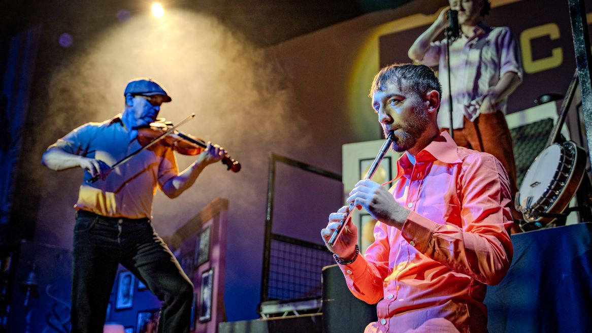 A man plays a pipe instrument while a man plays the fiddle and another stands in front of microphone