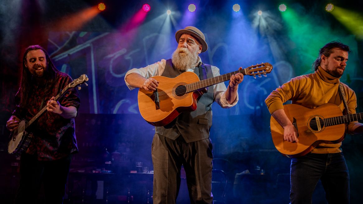 An old man with a large white beard plays guitar with two younger men